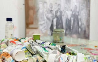 A close-up of an artist's workspace with oil paint tubes, brushes, and a jar of mixed paint, alongside an unfinished artwork featuring a historical scene, showcasing the process of professional art creation, international shipping available.