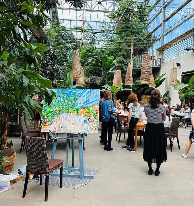 Art exhibition in a greenhouse with visitors viewing a colorful landscape painting by Nikolaus Kriese, surrounded by lush greenery and hanging woven lamps.